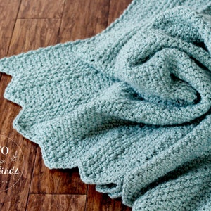 May include: A soft, light blue crocheted blanket with a scalloped edge. The blanket is folded and draped over a wooden surface. The text "TWO little birds patterns" is visible in the lower left corner of the image.
