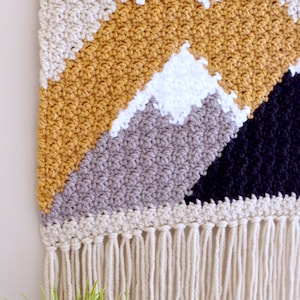 May include: A crocheted wall hanging with a geometric mountain design in shades of beige, mustard yellow, gray, white, and black. The hanging has a fringe border.