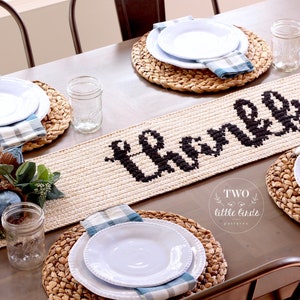 May include: A crocheted table runner with the word "thankful" in black yarn on a beige background. The runner is displayed on a wooden table with white plates, blue napkins, and woven placemats.