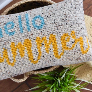 May include: A crocheted pillow with the words "hello summer" in blue and yellow lettering on a white background with speckles of color.