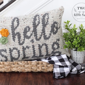 May include: A crocheted pillow with the words "Hello Spring" in gray yarn on a white background. The pillow has a floral embellishment with white, orange, and green yarn. The pillow is sitting in a woven basket with a black and white checkered fabric.