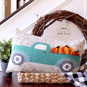 May include: A crocheted cushion featuring a teal truck carrying pumpkins. The cushion is placed on a wicker basket and a black and white checked fabric. The text "TWO little birds patterns" is on a brown wreath behind the cushion.