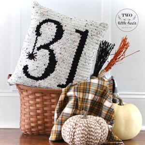May include: A white crocheted pillow with black yarn forming the numbers "31" and a small black spider with red eyes. The pillow is sitting in a brown wicker basket with a plaid blanket and two pumpkins.