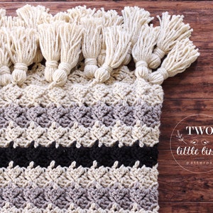 Peut inclure: Une couverture au crochet faite à la main avec des pompons crème sur le bord supérieur. La couverture présente un motif texturé avec des rayures crème, grises et noires. Le texte "TWO little birds patterns" est visible.