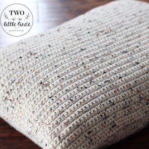 May include: A close-up of a cream-colored crocheted pillow with a speckled pattern. The pillow is made with a chunky yarn and has a textured surface.