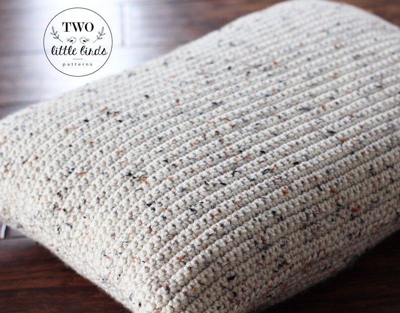 May include: A close-up of a cream-colored crocheted pillow with a speckled pattern. The pillow is made with a chunky yarn and has a textured surface.