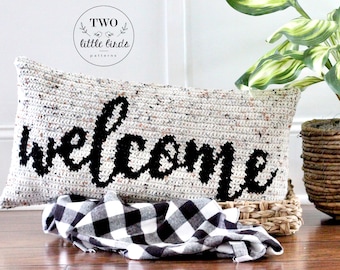 May include: A white crocheted pillow with the word "welcome" in black script. The pillow is sitting on a black and white checkered throw blanket and a woven basket.