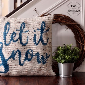 K&ouml;nnte beinhalten: Ein geh&auml;keltes Kissen mit dem Text "Let it snow" in blauer Wolle auf wei&szlig;em Hintergrund. Das Kissen liegt auf einem Holzboden mit einer kleinen Topfpflanze und einem Kranz im Hintergrund.