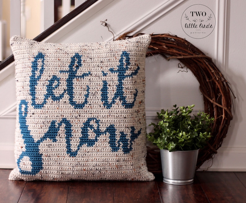 K&ouml;nnte beinhalten: Ein geh&auml;keltes Kissen mit dem Text "Let it snow" in blauer Wolle auf wei&szlig;em Hintergrund. Das Kissen liegt auf einem Holzboden mit einer kleinen Topfpflanze und einem Kranz im Hintergrund.