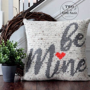 Pu&ograve; includere: Un cuscino bianco all'uncinetto con le parole "Be Mine" in filato grigio e un cuore rosso al centro. Il cuscino &egrave; appoggiato su un pavimento di legno con una ghirlanda e una pianta in vaso sullo sfondo.