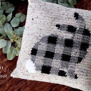 May include: A crocheted pillow with a black and white buffalo plaid design in the shape of a bunny. The pillow is on a wooden surface with green foliage.