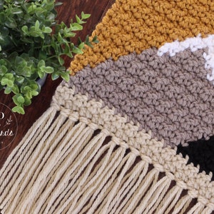 May include: A crocheted wall hanging with a geometric pattern in shades of mustard yellow, beige, gray, white, and black. The hanging has a fringe of natural colored yarn.