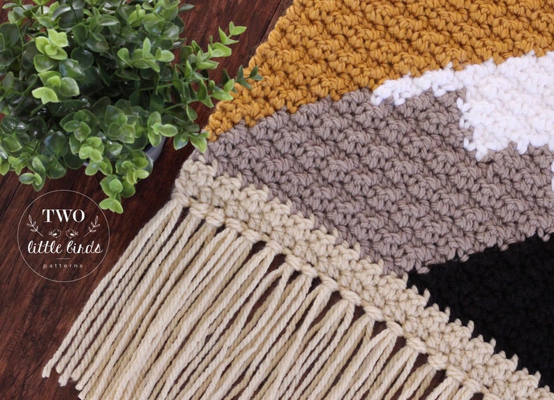 May include: A crocheted wall hanging with a geometric pattern in shades of mustard yellow, beige, gray, white, and black. The hanging has a fringe of natural colored yarn.