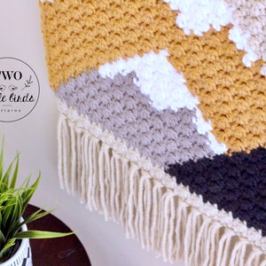 May include: A geometric wall hanging with a fringe border. The hanging is made of crocheted yarn in shades of mustard yellow, white, gray, and black.