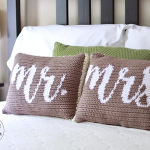 Op de afbeelding: Twee bruine gehaakte kussens met witte letters. De kussens zeggen "Mr." en "Mrs." en liggen op een wit bed.