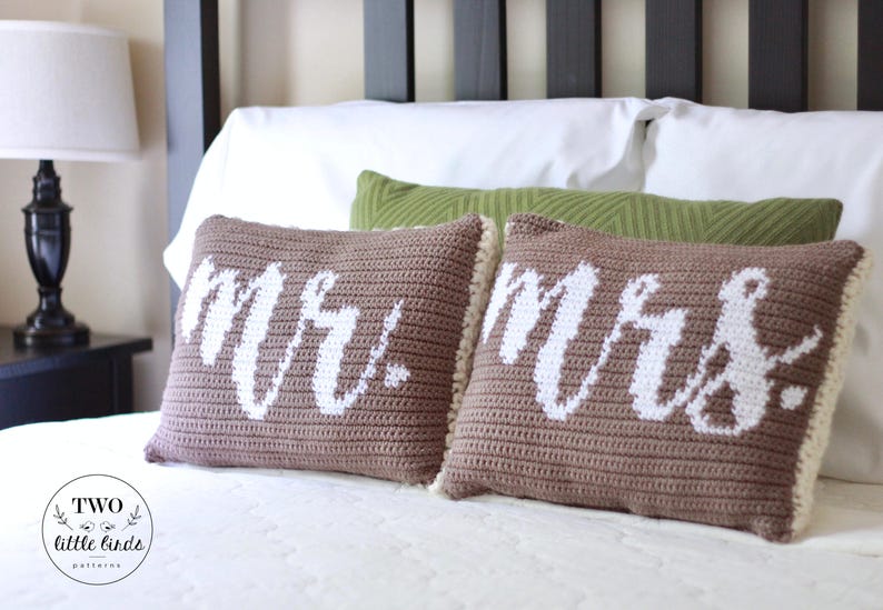 Op de afbeelding: Twee bruine gehaakte kussens met witte letters. De kussens zeggen "Mr." en "Mrs." en liggen op een wit bed.