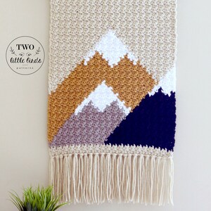 May include: A beige crocheted wall hanging with a mountain scene. The mountains are made of different colored yarn, including mustard yellow, gray, and navy blue. The wall hanging has a fringe border.