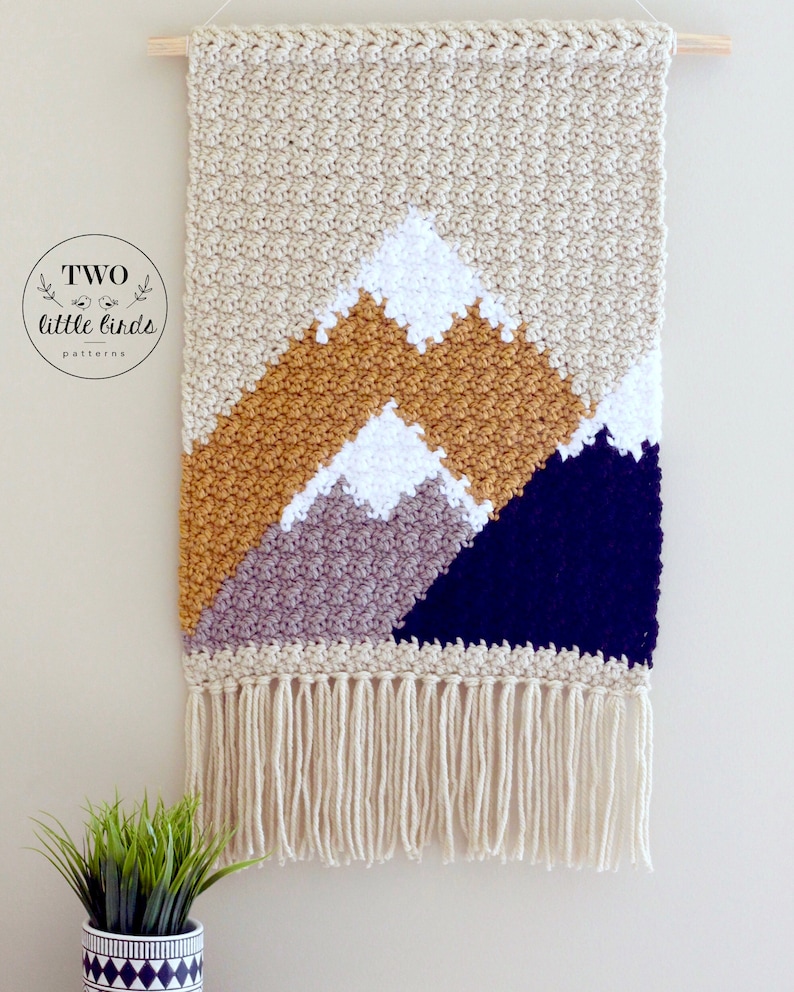 May include: A beige crocheted wall hanging with a mountain scene. The mountains are made of different colored yarn, including mustard yellow, gray, and navy blue. The wall hanging has a fringe border.