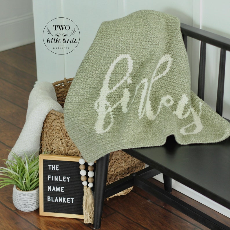 May include: A sage green crocheted blanket with the name "Finley" in white script. The blanket is draped over a black bench. A sign reads "The Finley Name Blanket" next to a basket and a small plant.