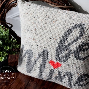 Pu&ograve; includere: Un cuscino bianco all'uncinetto con un design grigio "be mine" e un cuore rosso. Il cuscino &egrave; su una superficie di legno.