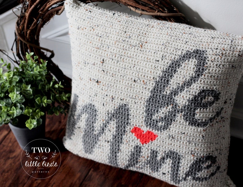 Pu&ograve; includere: Un cuscino bianco all'uncinetto con un design grigio "be mine" e un cuore rosso. Il cuscino &egrave; su una superficie di legno.