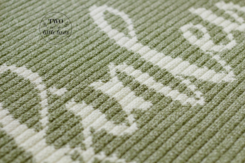 May include: Close-up of a textured, olive green and white fabric with a repeating pattern. The fabric has a ribbed texture with a white design. The words "TWO little birds" are in a circle in the upper left corner.