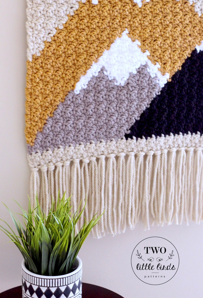 May include: A crocheted wall hanging with a geometric mountain design in shades of beige, mustard yellow, gray, white, and black. The hanging has a fringe border.