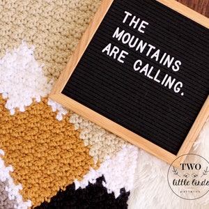 May include: A crochet blanket with a geometric pattern in shades of beige, white, mustard yellow, gray, and black. A letter board with the text "THE MOUNTAINS ARE CALLING." is on top of the blanket. The letter board is made of black felt with a wooden frame.