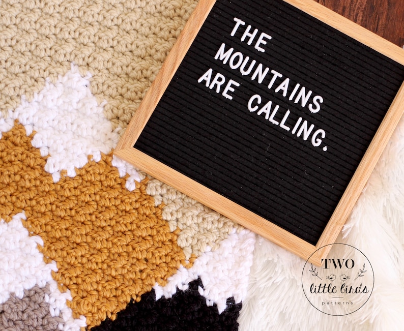 May include: A crochet blanket with a geometric pattern in shades of beige, white, mustard yellow, gray, and black. A letter board with the text "THE MOUNTAINS ARE CALLING." is on top of the blanket. The letter board is made of black felt with a wooden frame.