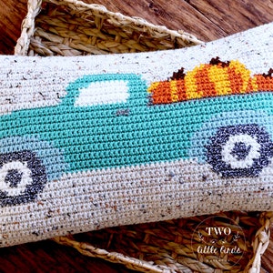 May include: A crocheted cushion featuring a teal blue truck with a white windscreen and black wheels. The truck is carrying a stack of orange and yellow pumpkins. The cushion is made with a beige speckled yarn.