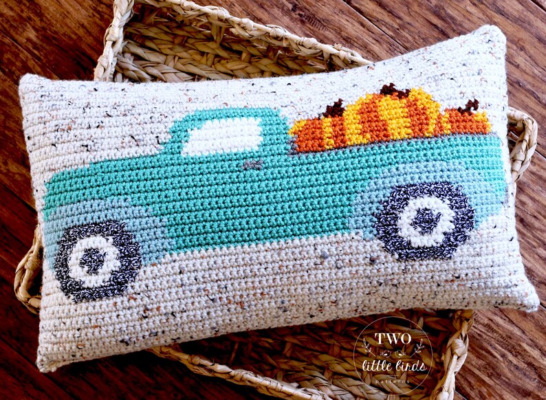 May include: A crocheted cushion featuring a teal blue truck with a white windscreen and black wheels. The truck is carrying a stack of orange and yellow pumpkins. The cushion is made with a beige speckled yarn.
