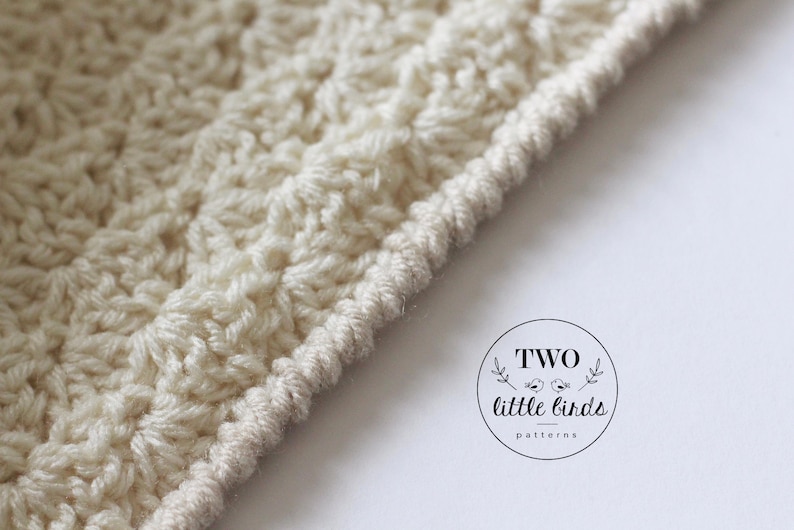 May include: Close-up of a cream-colored crocheted fabric with a textured stitch pattern. The fabric has a finished edge with a decorative border.