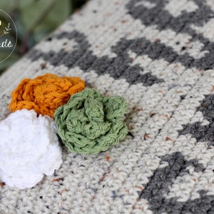 May include: A close-up of a crocheted throw pillow with a geometric pattern in shades of gray and white. Three crocheted flowers in white, green, and orange are attached to the pillow.