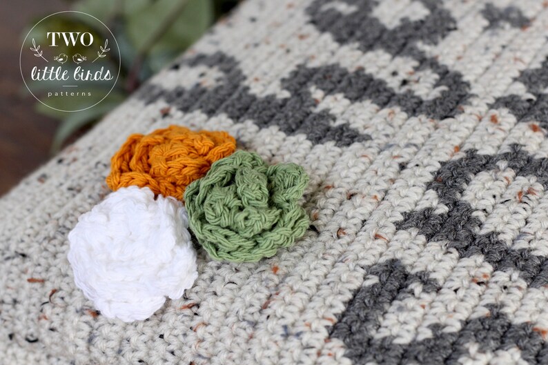 May include: A close-up of a crocheted throw pillow with a geometric pattern in shades of gray and white. Three crocheted flowers in white, green, and orange are attached to the pillow.