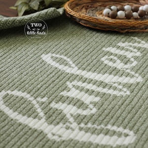 May include: A sage green knitted blanket with the word "hello" in white, ribbed texture. The blanket is folded and displayed with a woven basket and decorative beads in the background. The blanket has the text "two little birds" in a circle.