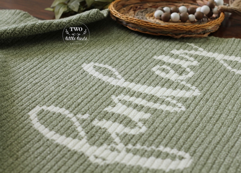 May include: A sage green knitted blanket with the word "hello" in white, ribbed texture. The blanket is folded and displayed with a woven basket and decorative beads in the background. The blanket has the text "two little birds" in a circle.