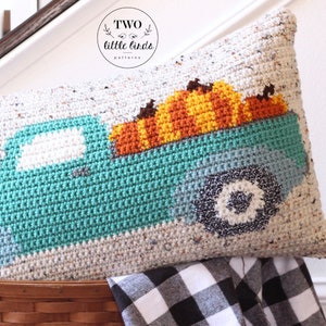 May include: A crocheted cushion featuring a teal truck carrying a pile of orange, yellow, and brown pumpkins. The cushion is made with a speckled white yarn and has a black and white wheel.