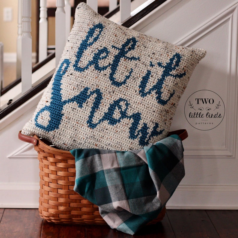 K&ouml;nnte beinhalten: Ein wei&szlig;es und blaues H&auml;kelkissen mit dem Text "Let it snow" in Blau. Das Kissen liegt in einem braunen Weidenkorb mit einer gr&uuml;n-wei&szlig; karierten Decke.