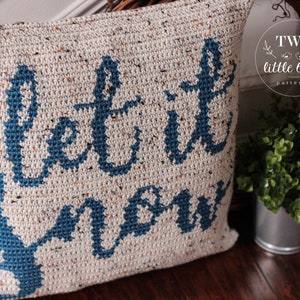 K&ouml;nnte beinhalten: Ein wei&szlig;es H&auml;kelkissen mit den Worten "Let it snow" in blauer Schrift. Das Kissen hat eine gesprenkelte Textur und ist mit einem blauen Anker verziert.
