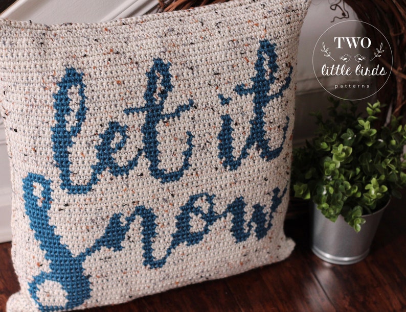 K&ouml;nnte beinhalten: Ein wei&szlig;es H&auml;kelkissen mit den Worten "Let it snow" in blauer Schrift. Das Kissen hat eine gesprenkelte Textur und ist mit einem blauen Anker verziert.