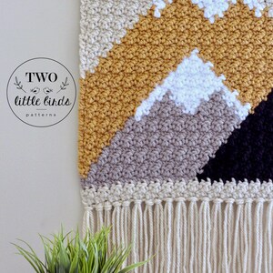 May include: A crocheted wall hanging with a mountain design in beige, mustard yellow, gray, white, and black. The hanging has a fringe border.