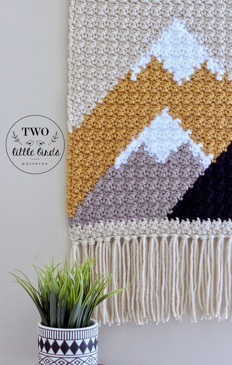 May include: A crocheted wall hanging with a mountain design in beige, mustard yellow, gray, white, and black. The hanging has a fringe border.