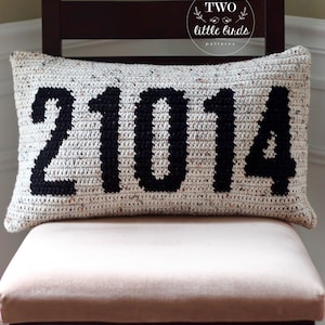 May include: A white crocheted cushion with black yarn forming the numbers "21014". The cushion is on a light brown chair with a dark brown wooden frame. The cushion has a tag that reads "TWO little birds patterns".