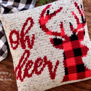 May include: A crocheted pillow with a red and black buffalo plaid deer silhouette and the text "Oh Deer" in red. The pillow is on a wooden surface.