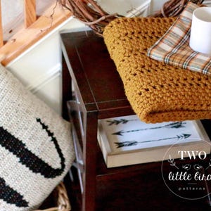 Pu&ograve; includere: Una coperta in crochet di colore giallo senape con un tessuto a quadri sottostante. La coperta &egrave; drappeggiata su un tavolino di legno con una tazza bianca e un cuscino con una lettera W nera. Il tavolino ha un cartello che dice "Two Little Birds Patterns".