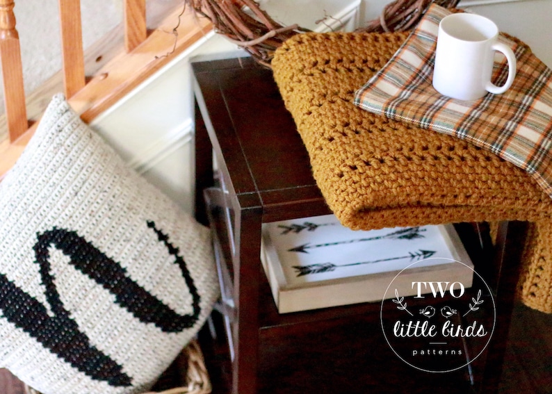 Pu&ograve; includere: Una coperta in crochet di colore giallo senape con un tessuto a quadri sottostante. La coperta &egrave; drappeggiata su un tavolino di legno con una tazza bianca e un cuscino con una lettera W nera. Il tavolino ha un cartello che dice "Two Little Birds Patterns".