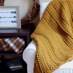Pu&ograve; includere: Una coperta in crochet color giallo senape drappeggiata su una poltrona bianca. La coperta ha un semplice motivo a punto ed &egrave; realizzata con un filato morbido.