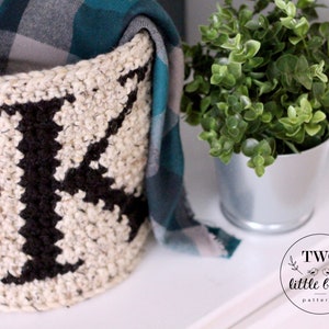 Op de afbeelding: Een witte gehaakte opbergmand met een zwarte letter "K" die in het ontwerp is geweven. De mand is gevuld met een geruite deken en staat op een wit oppervlak naast een kleine plant in een pot.
