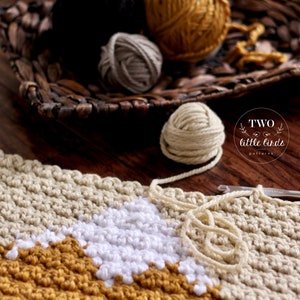 May include: A close-up of a crocheted blanket with a geometric pattern in white, beige, and mustard yellow. The blanket is being worked on with a crochet hook and yarn balls in various colors are visible in the background.