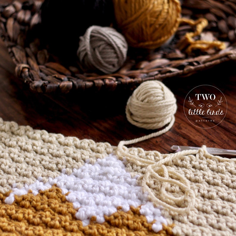 May include: A close-up of a crocheted blanket with a geometric pattern in white, beige, and mustard yellow. The blanket is being worked on with a crochet hook and yarn balls in various colors are visible in the background.
